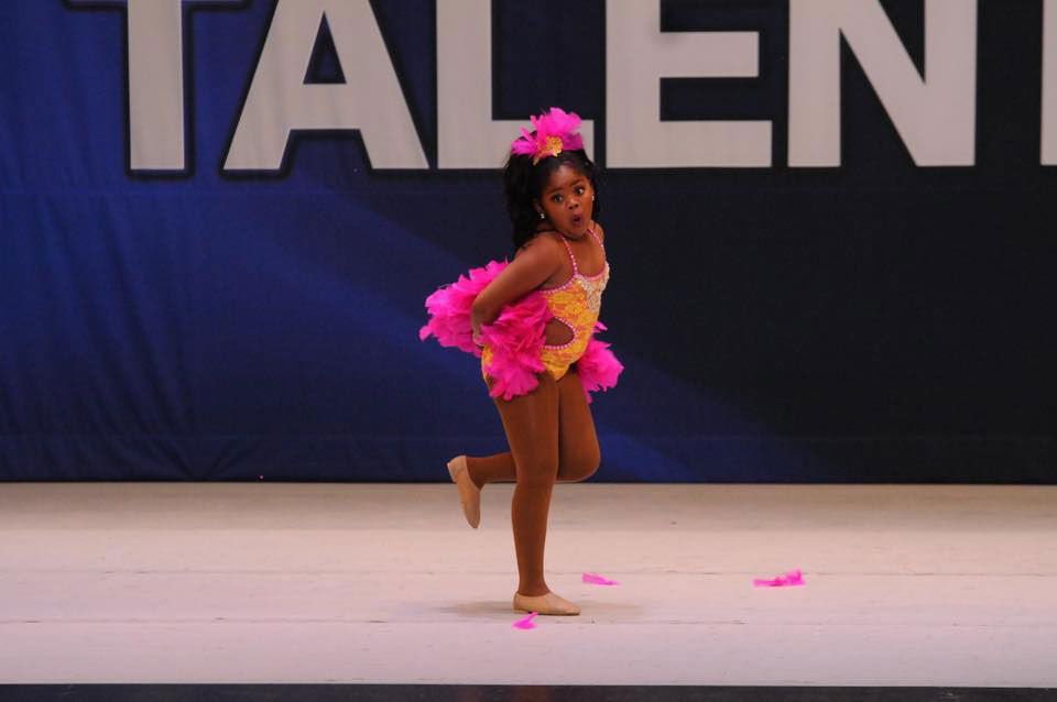 Young dancer in a pink and yellow costume performs on stage, smiling. Stage background reads