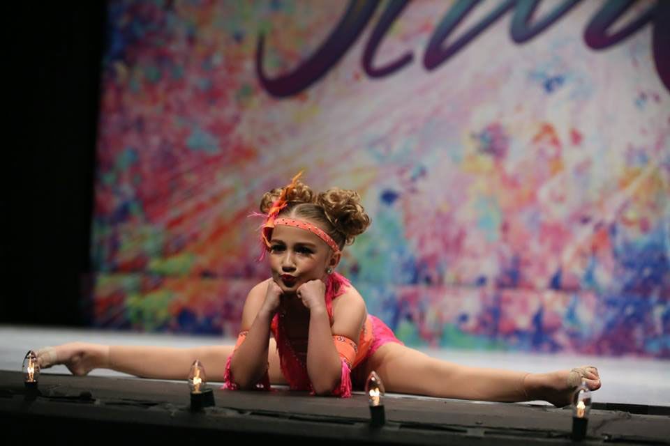 Young dancer in a split pose on stage, wearing pink and orange costume, looking at the camera.