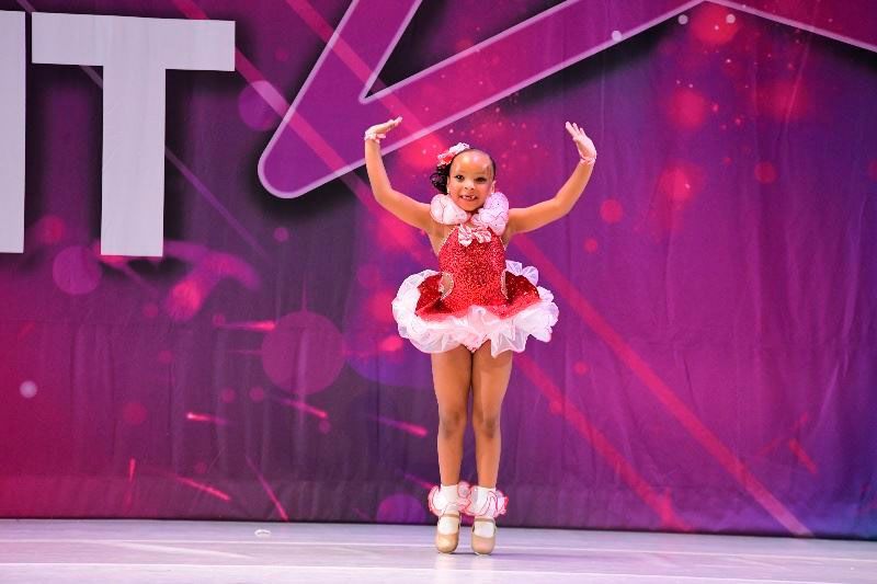 Young dancer in red and white costume on stage with arms raised.