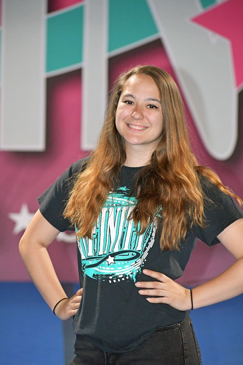 Teenage girl with long brown hair smiles, hands on hips, in front of a colorful backdrop.