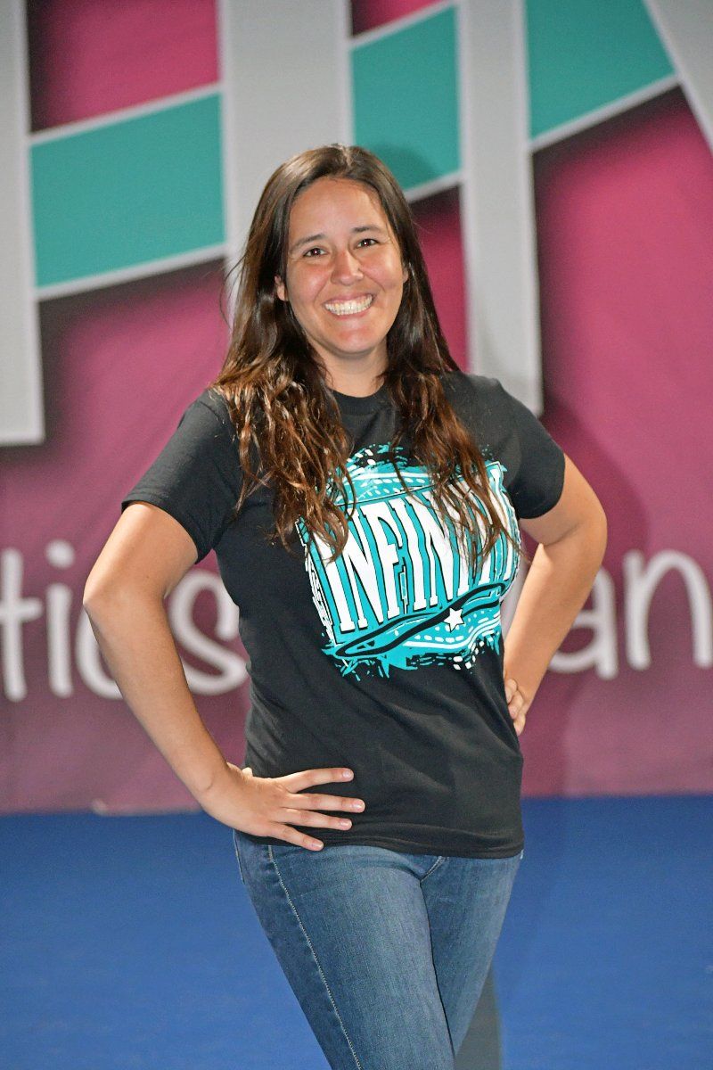 Woman with hands on hips, smiling, wearing a black tee and jeans, standing in a gym.