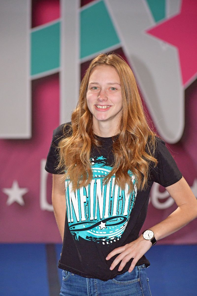 Teen girl with long hair, smiling, wearing a black t-shirt and jeans, posing indoors with a hand on her hip.