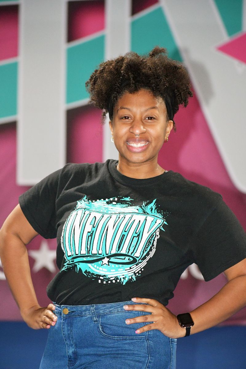 Woman with curly hair, arms on hips, wearing a black t-shirt and jeans. Background with colorful logo.