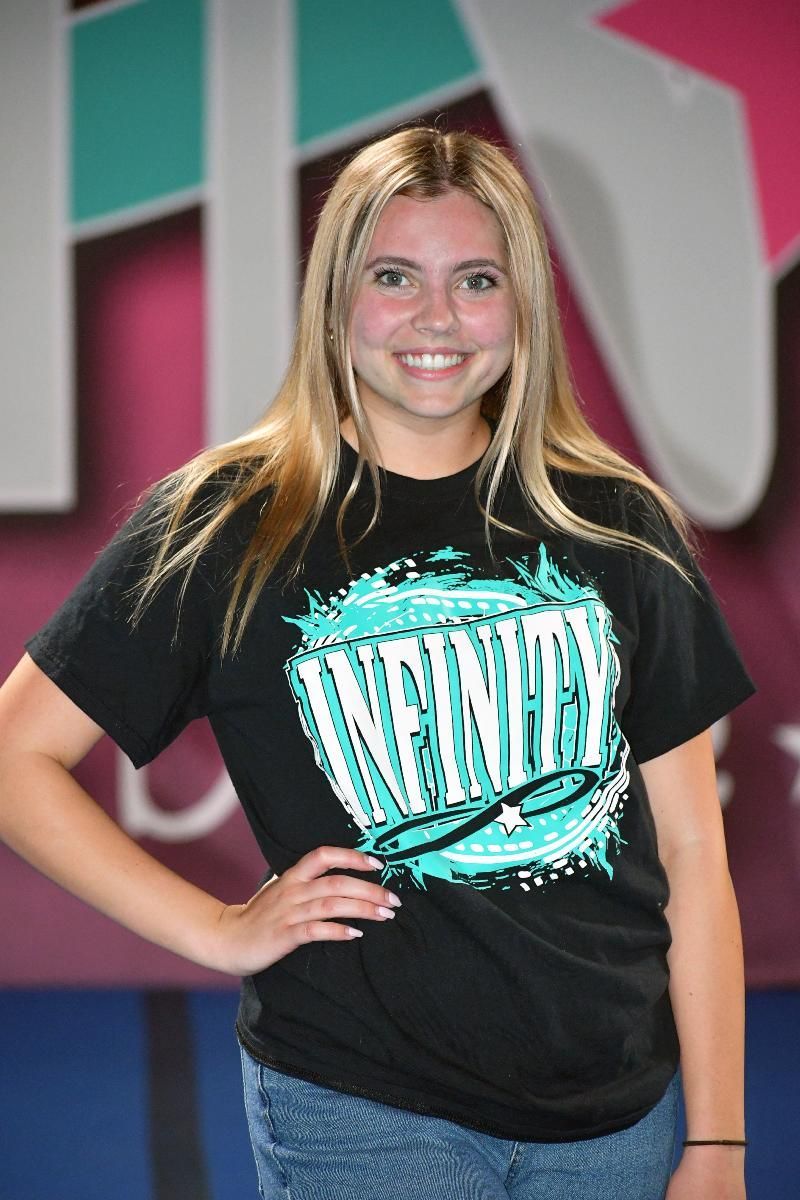 Blonde girl smiling, wearing black shirt with teal graphic, and jeans, in front of a dance studio backdrop.
