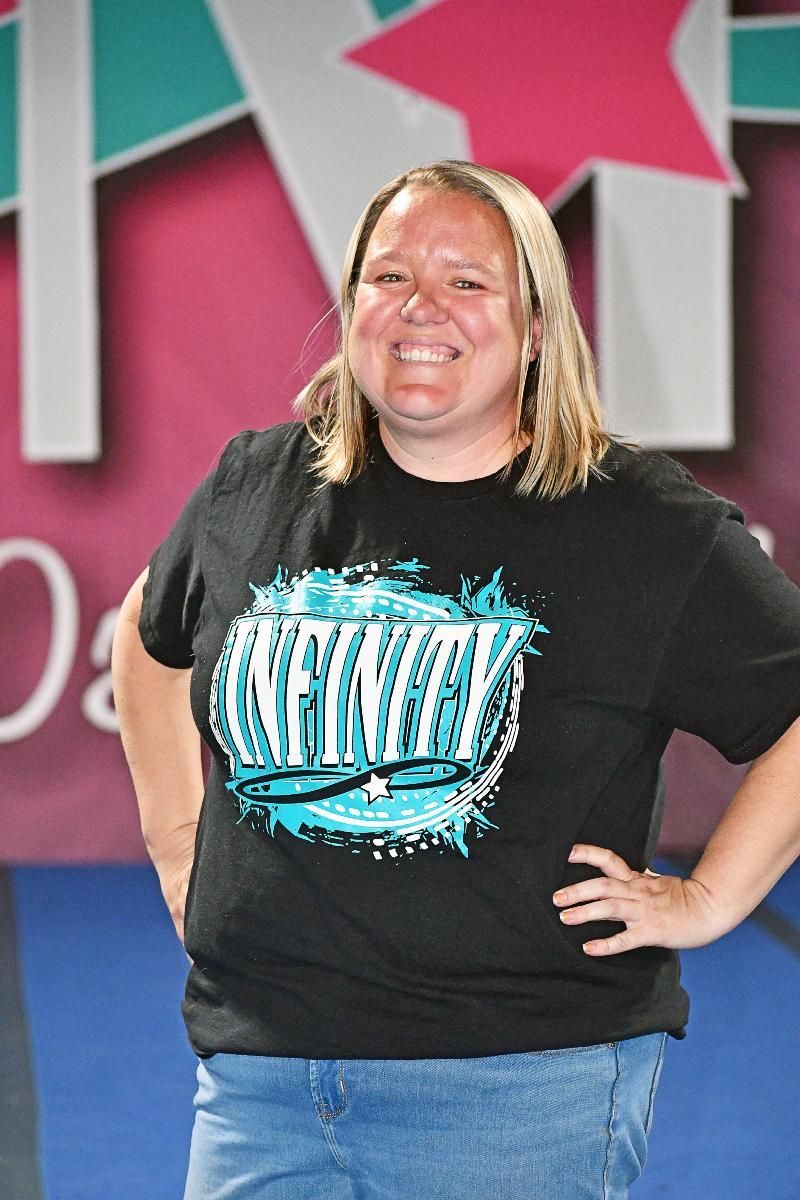 Woman in black t-shirt smiles, hands on hips. Light blonde hair, blue jeans. Stage backdrop.