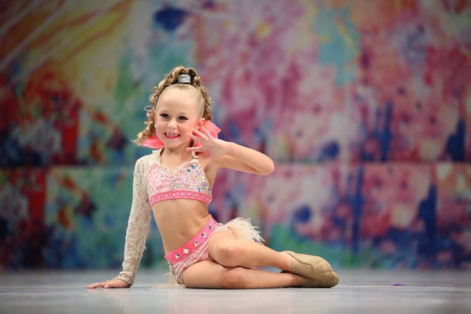 Young girl in pink dance costume waves, smiling, stage backdrop.