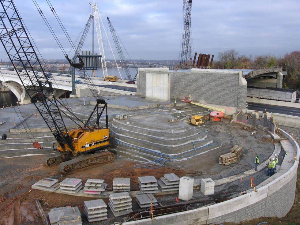 Construction site with cranes, concrete structures, and workers; bridge in background.