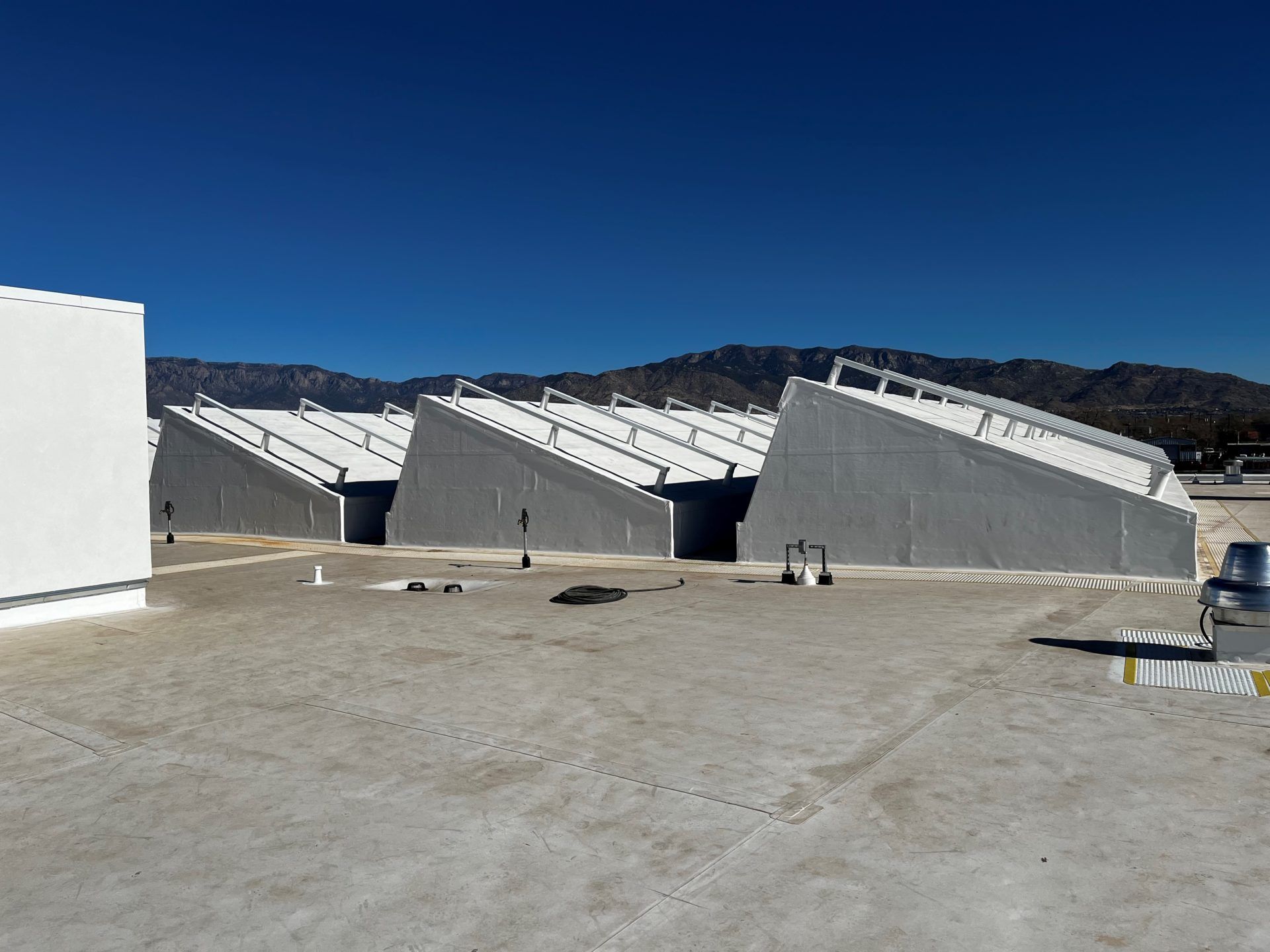 Three gray buildings with sloped roofs on a flat rooftop, mountains in the background, blue sky.