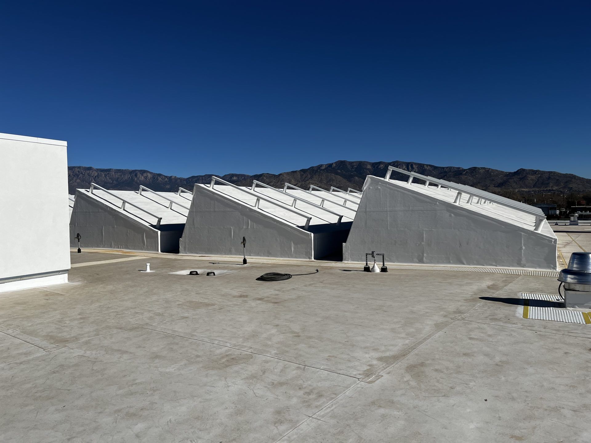 Gray industrial buildings with angled roofs on a flat rooftop against a backdrop of mountains and blue sky.