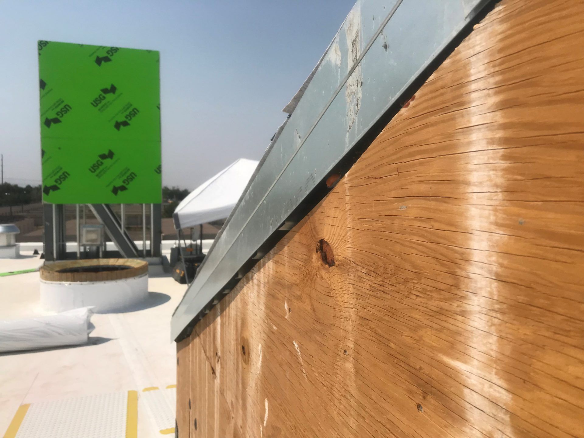 Wooden building exterior with metal trim against a sunny sky; a green protective sheet and equipment are visible in the background.