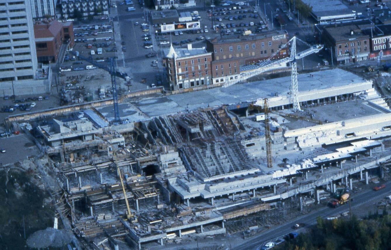 Construction site of a large building. Cranes and concrete structures dominate the scene. Urban setting with other buildings visible.