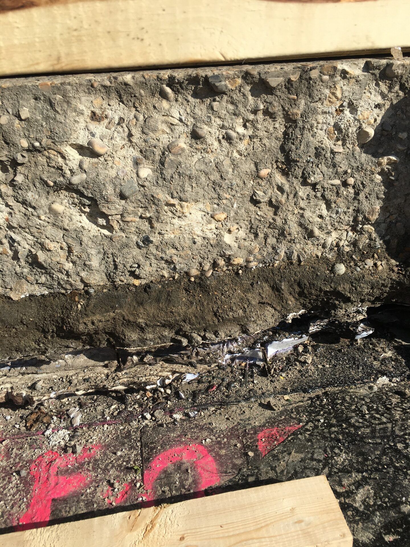 Cutaway of pavement showing layers of concrete, gravel, and asphalt with wooden boards and red markings.
