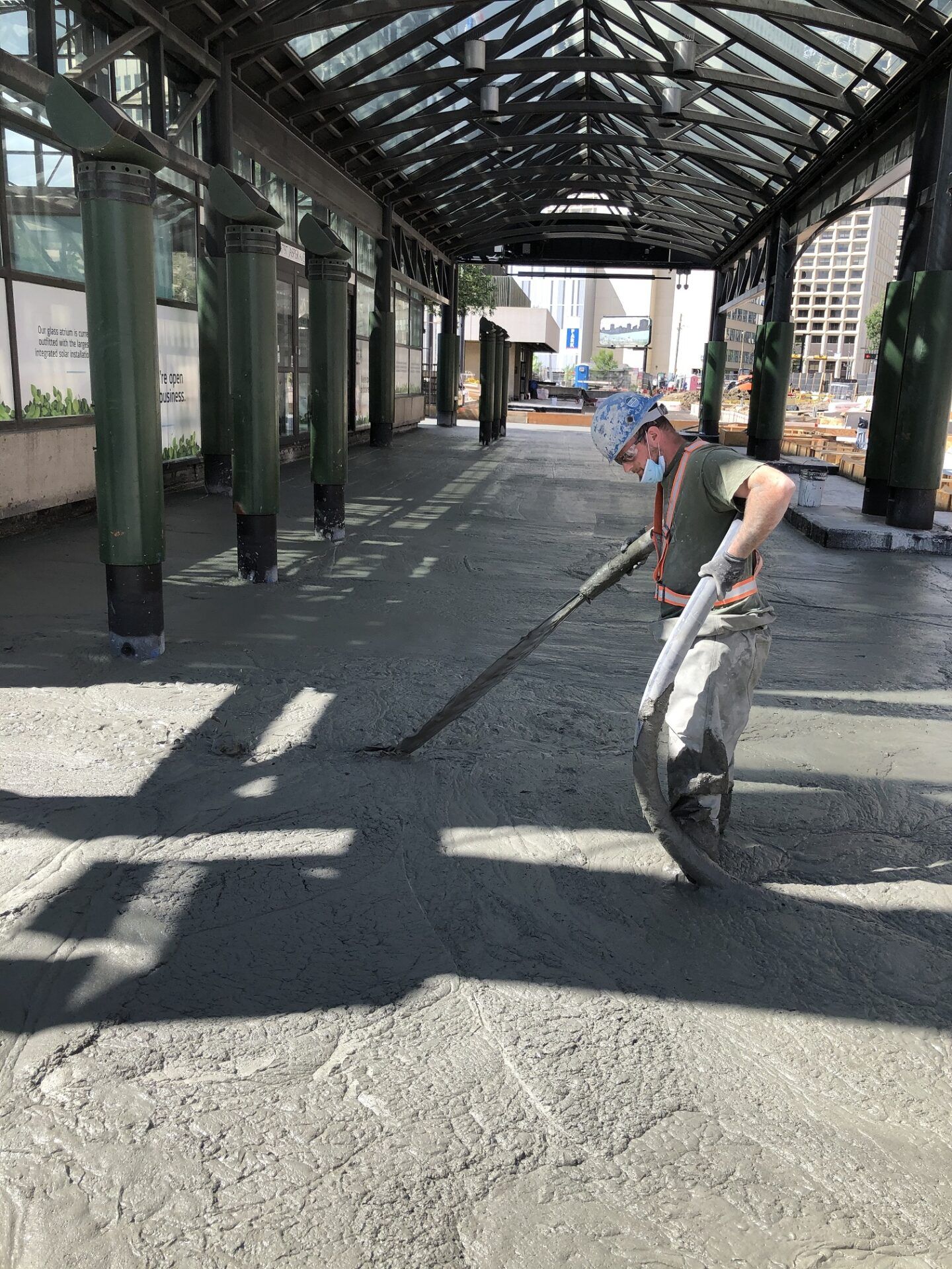 Man smoothing wet cement with a long-handled tool under a glass-roofed walkway.