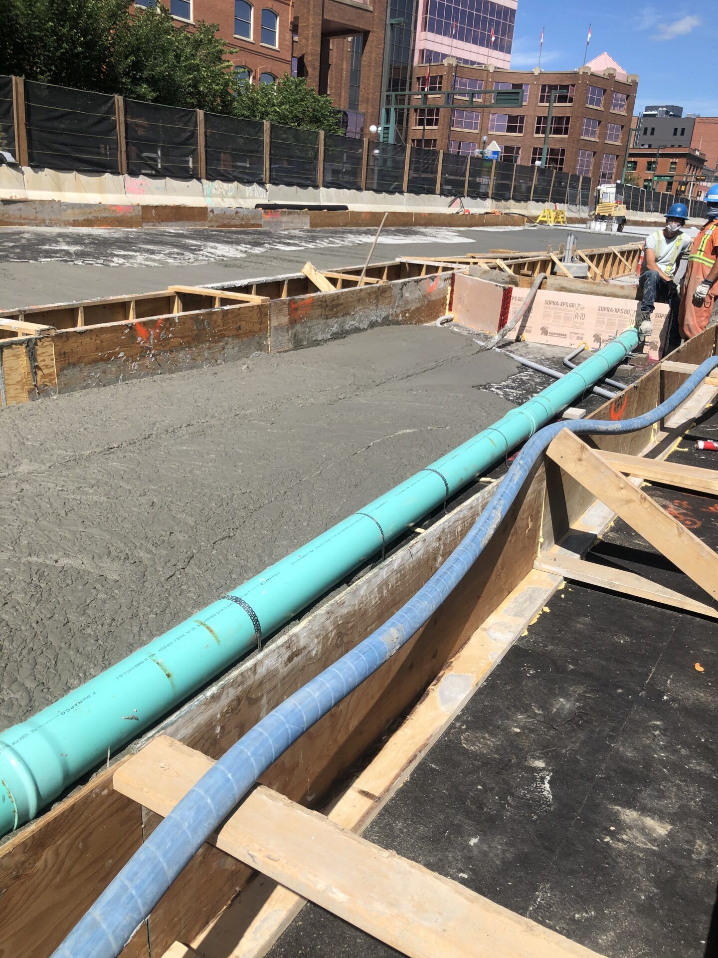 Construction site with concrete work. Green and blue pipes lie near fresh concrete, wooden forms, and workers in the background.