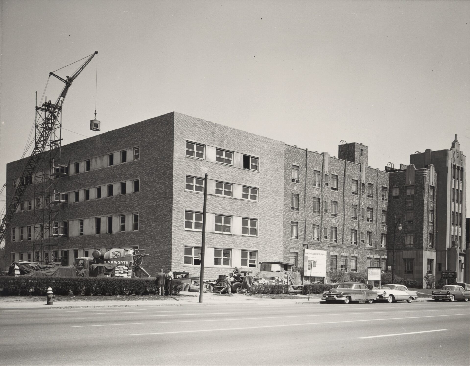 Construction site with brick building, crane, and cars on street.