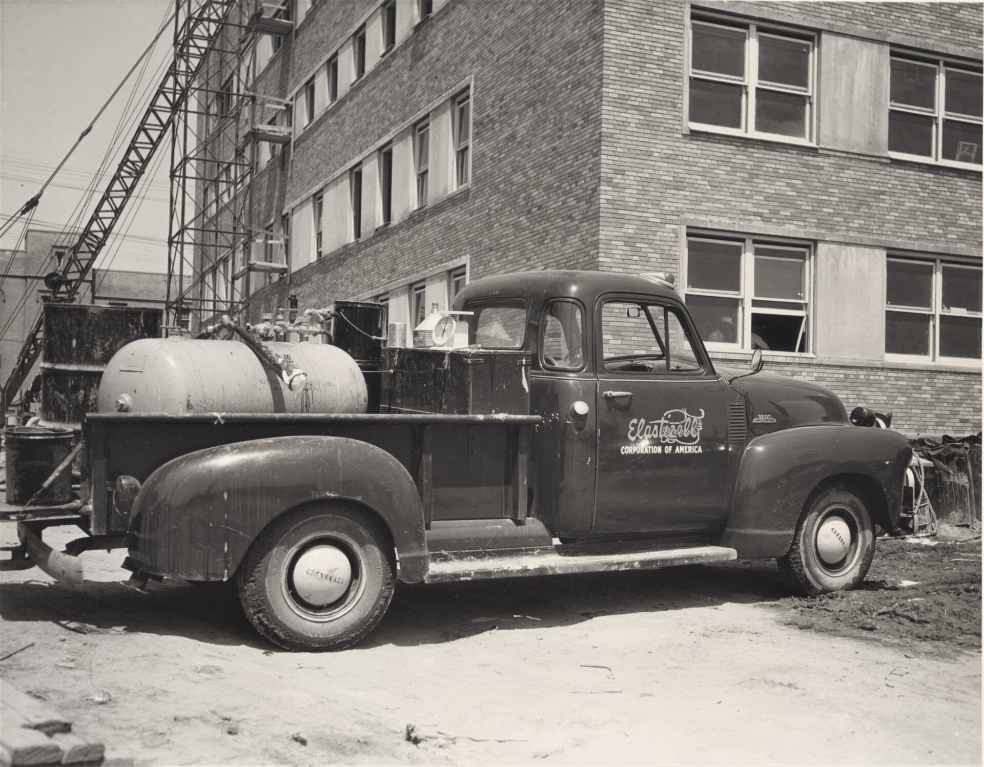 Vintage pickup truck with tank in bed parked near brick building under construction.