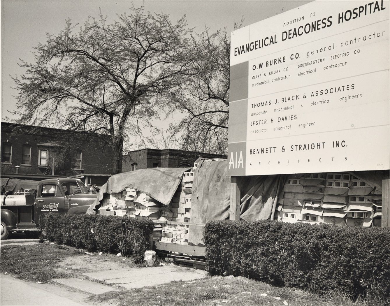 Black and white photo of a sign for Evangelical Deaconess Hospital next to construction materials. A truck is parked near a small building.