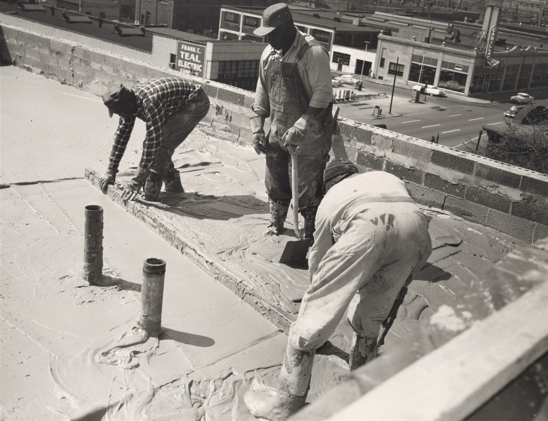 Three construction workers spread concrete on a flat rooftop, with city buildings visible in the background.
