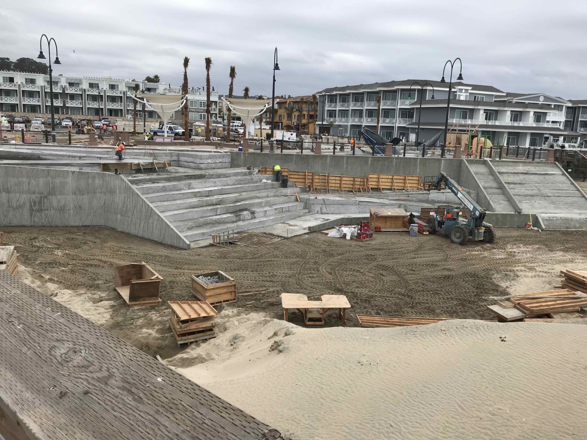 Construction site on a sandy beach featuring concrete stairs, machinery, and wooden forms with buildings in the background.