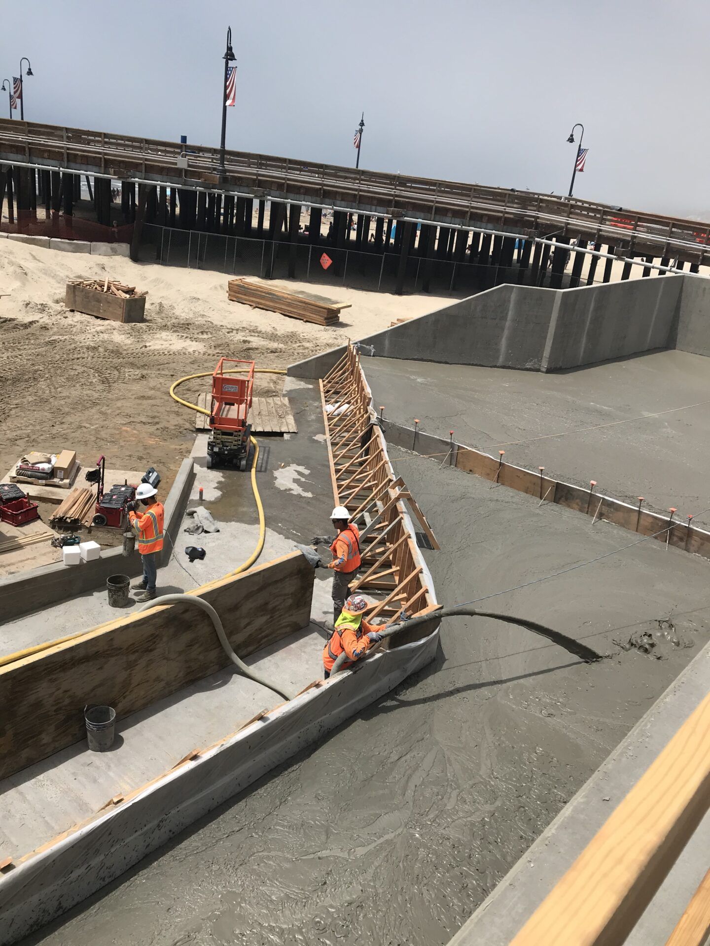 Construction workers pour concrete into wooden forms at an outdoor job site near a pier.