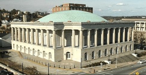 A stately, light-colored stone building with a colonnade and a large, teal, rounded dome under a clear sky.
