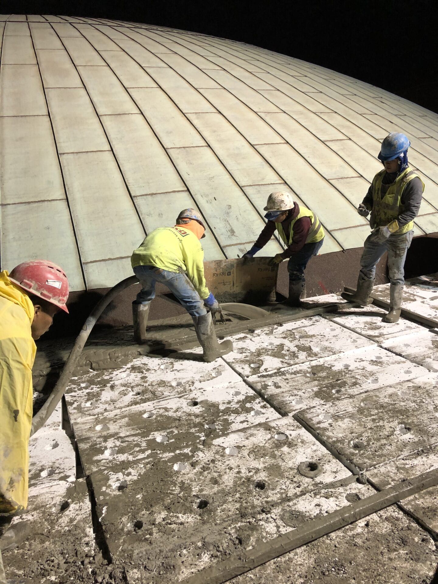 Construction workers in safety gear apply concrete to a curved structure under night lighting.