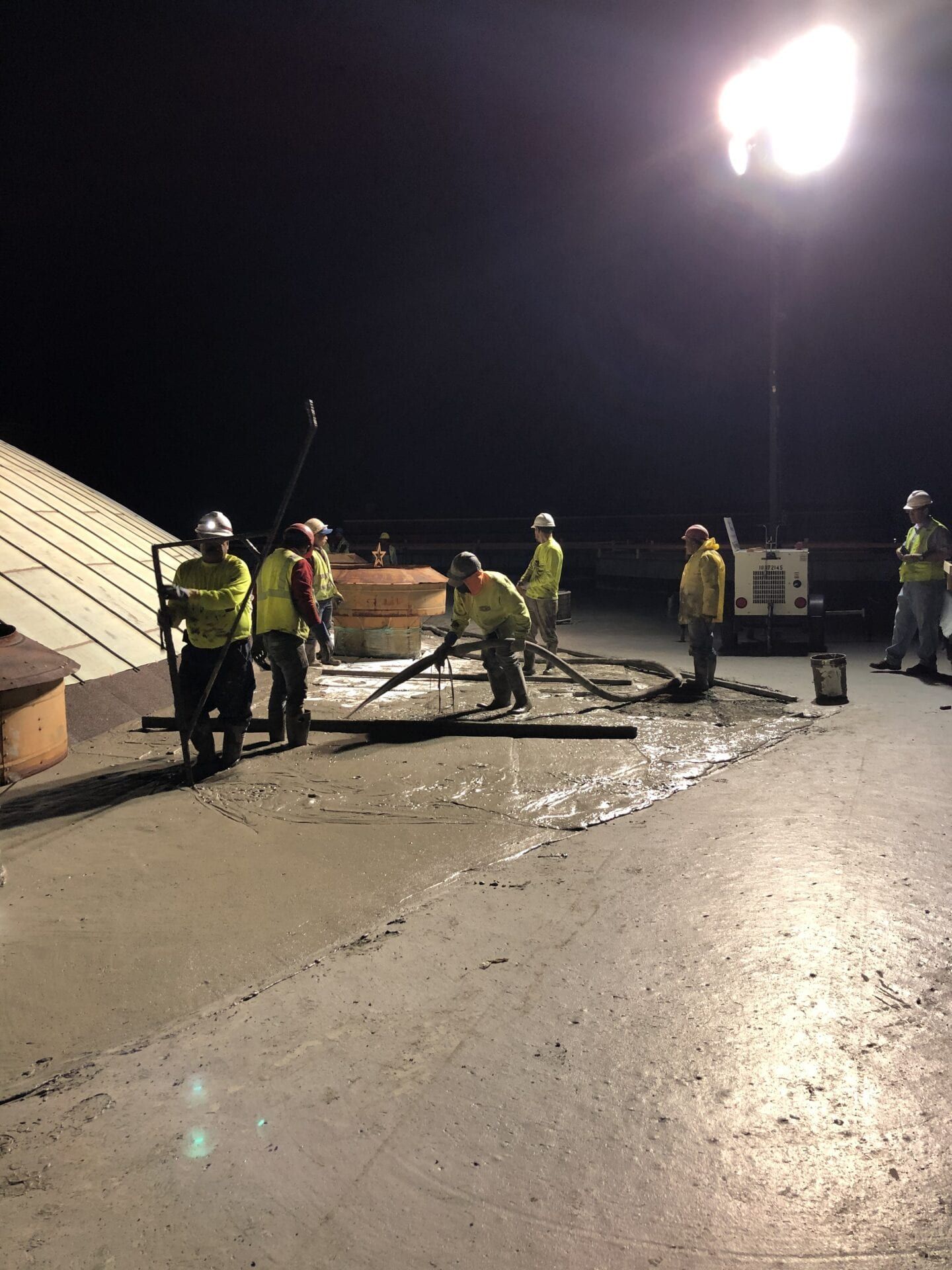 Workers in high-visibility gear work on a concrete surface under bright floodlights at night.