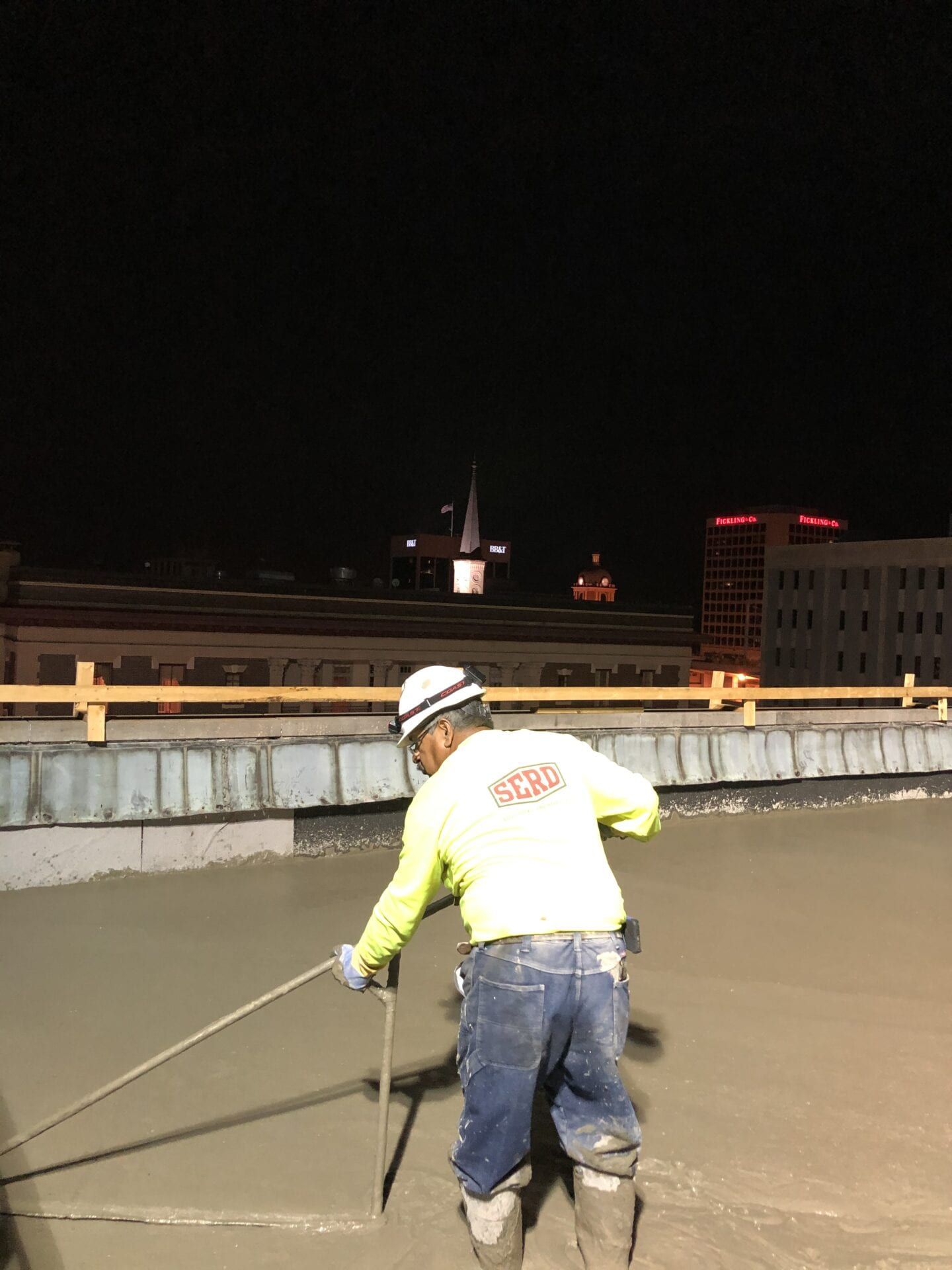 A worker in a high-visibility yellow shirt and hard hat uses a long-handled tool to smooth wet concrete on a rooftop at night.