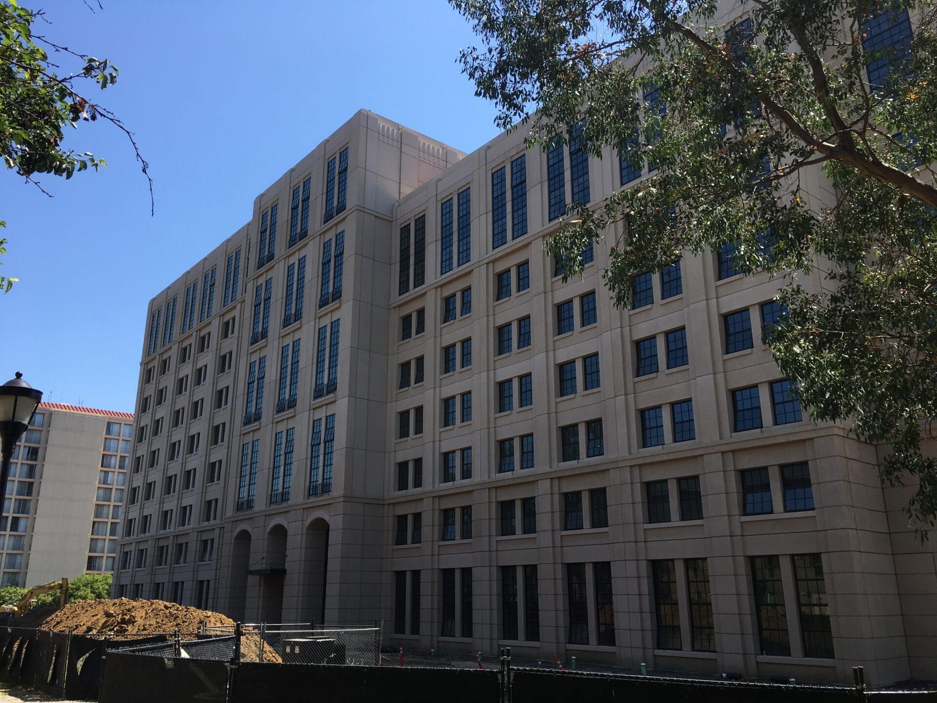 A large, multi-story beige building with rows of windows, partially framed by green trees against a clear blue sky.