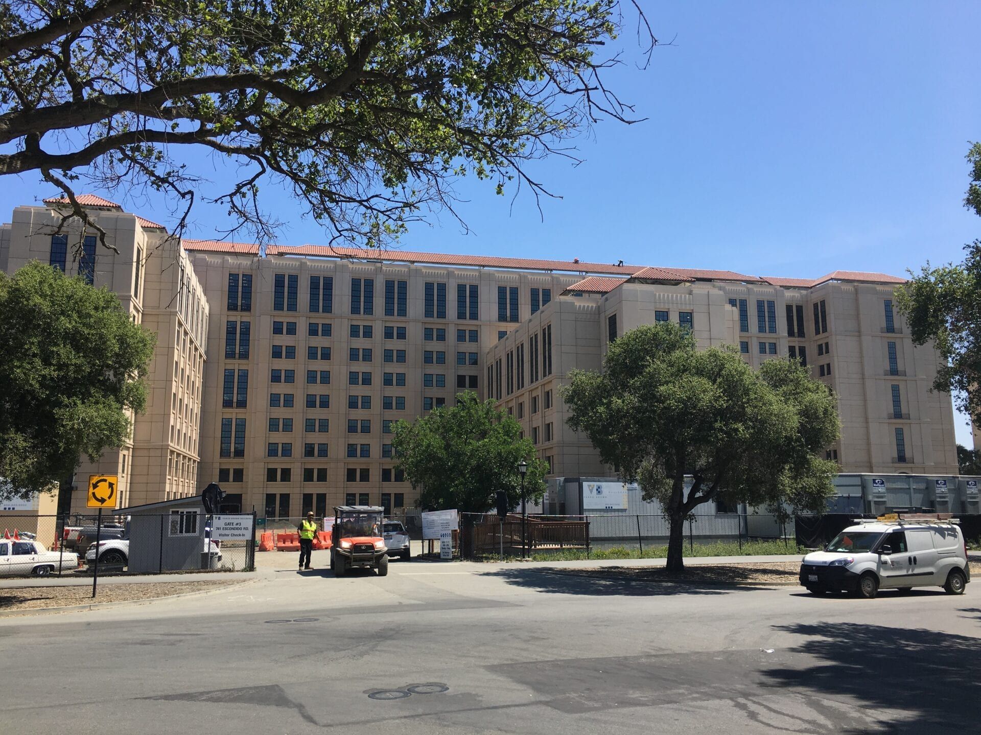 A large beige building under construction, viewed from a street with parked vehicles and mature trees under a clear sky.