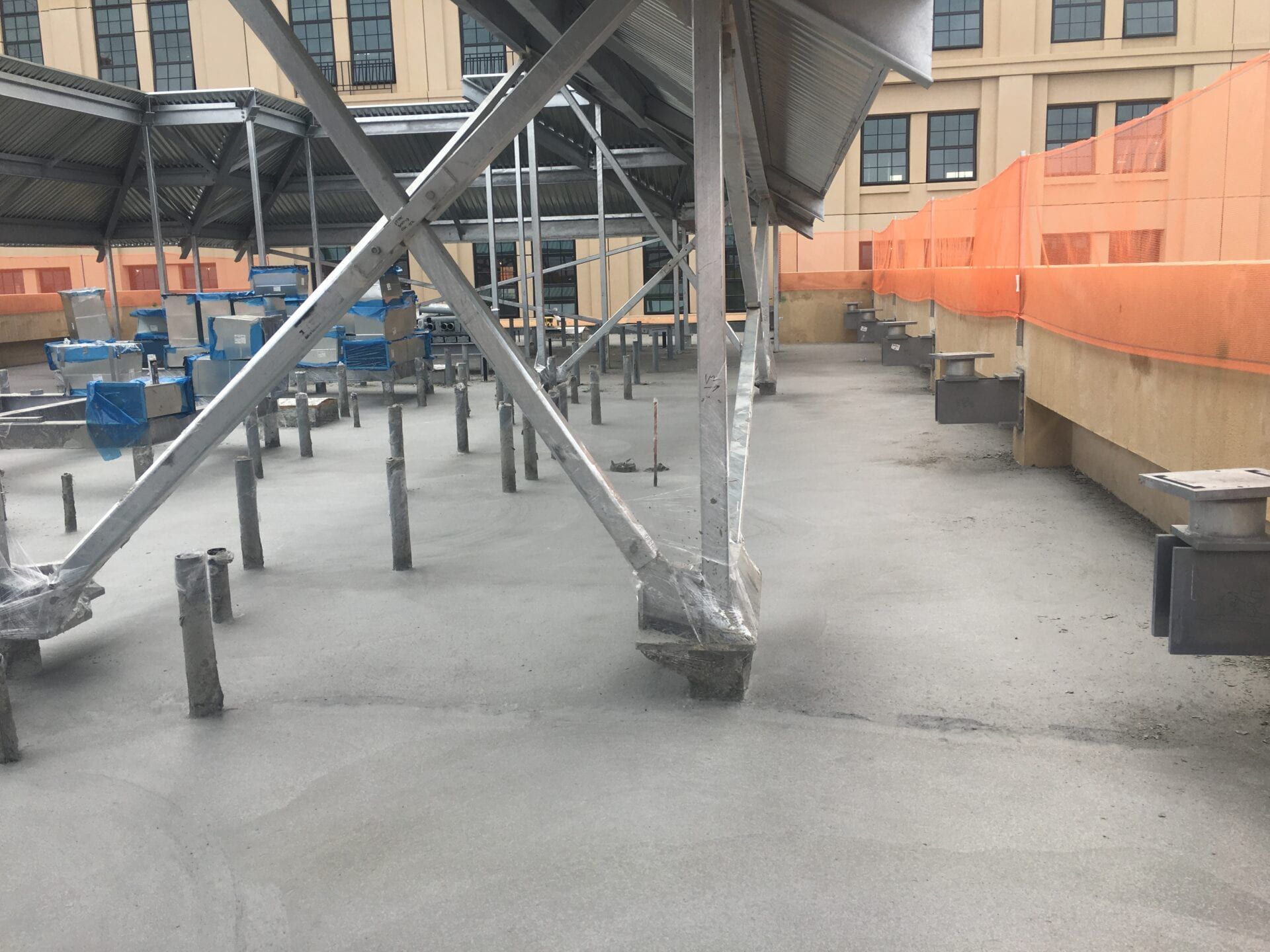 Construction site on a concrete rooftop with steel support beams, scaffolding, and orange safety fencing.