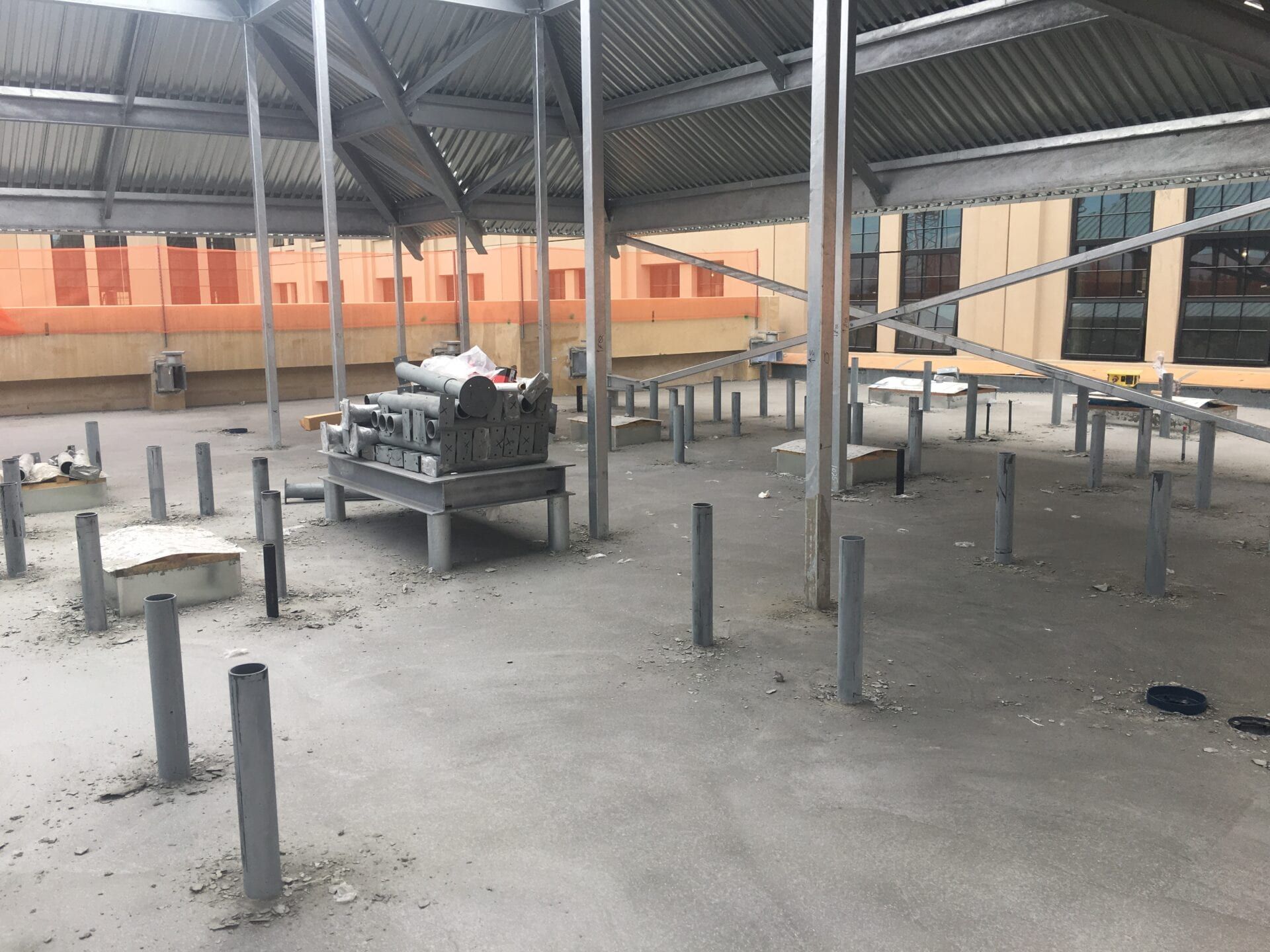 Construction site rooftop with metal support pillars, equipment, and a corrugated roof structure against a brick building.