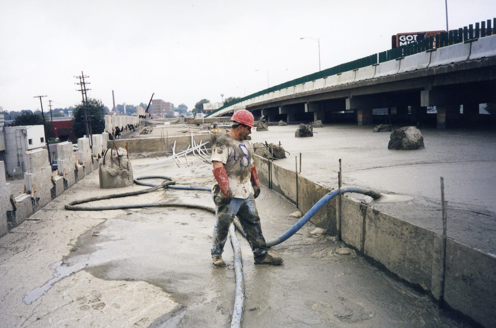 A worker in a red hard hat directs a hose on a concrete bridge surface during construction.