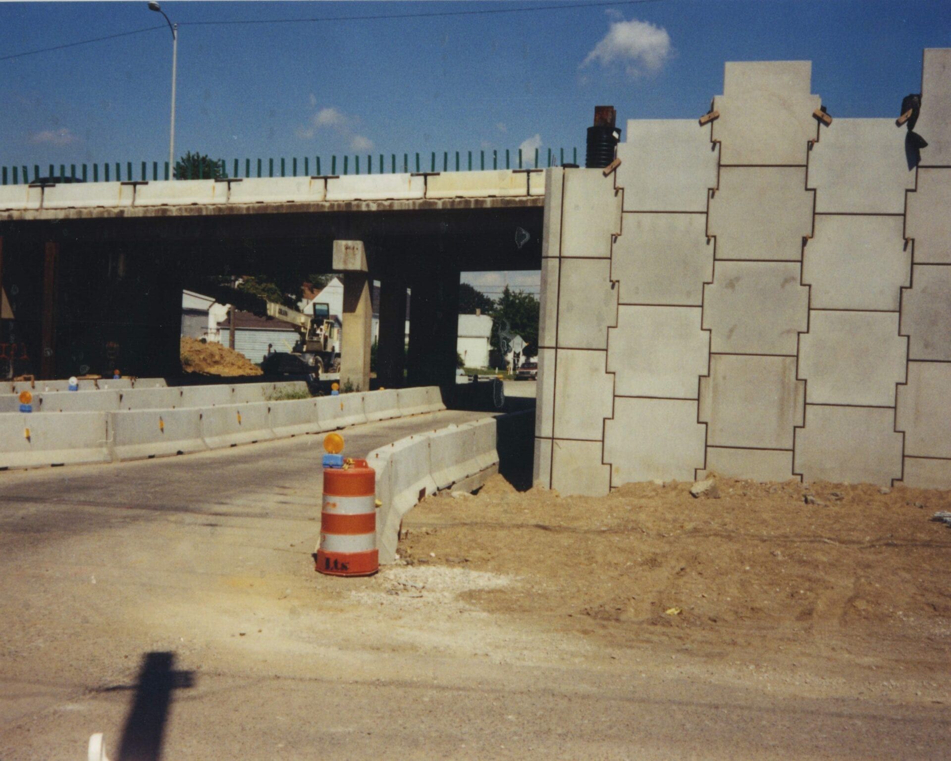 An underpass construction site with a tiered concrete retaining wall, a traffic barrel, and temporary barriers.