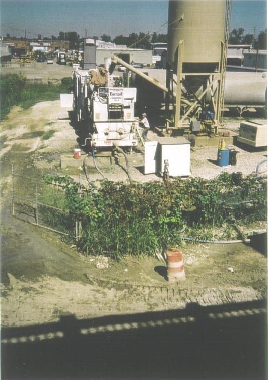 An industrial site with a tall storage silo, a mobile concrete mixer, and equipment positioned near a patch of greenery.