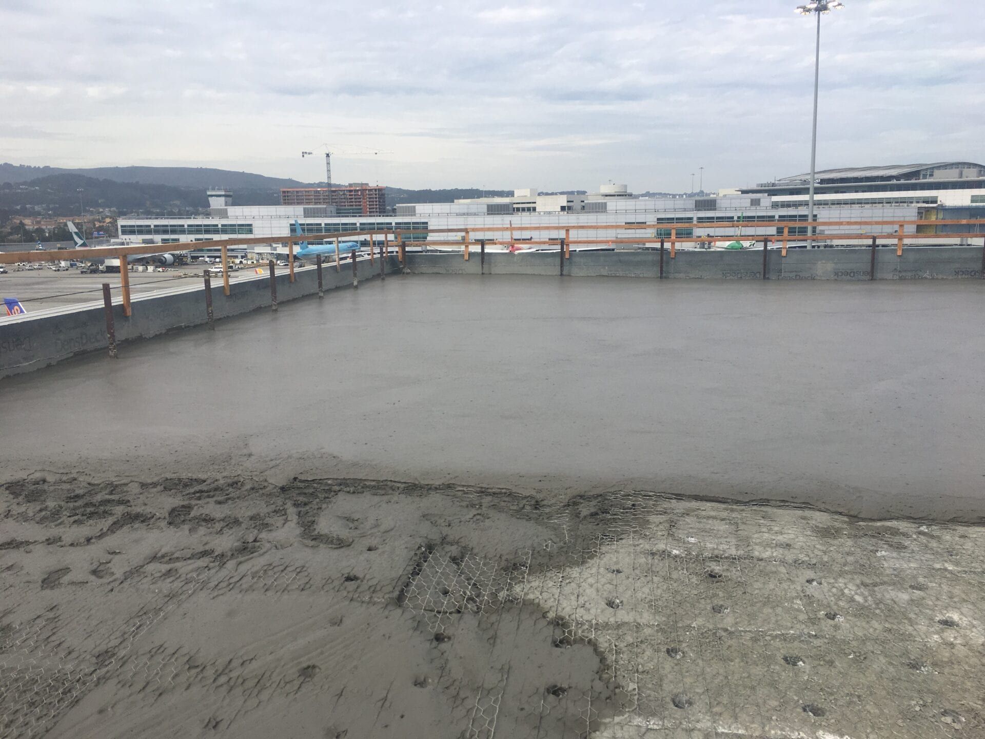 Freshly poured wet concrete covers a flat rooftop construction site, with a distant airport terminal in the background.