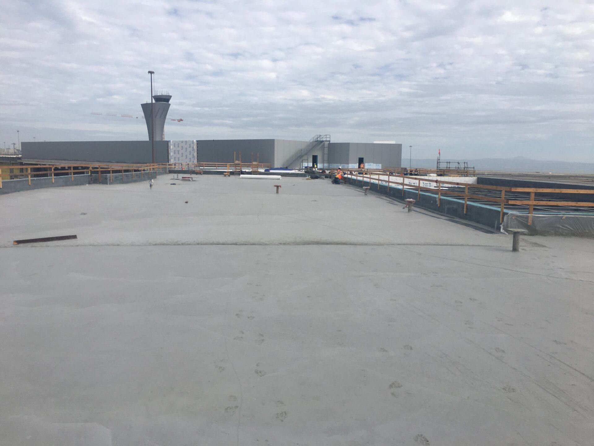 A wide, flat concrete rooftop under a cloudy sky, with a tall, modern airport control tower visible in the background.