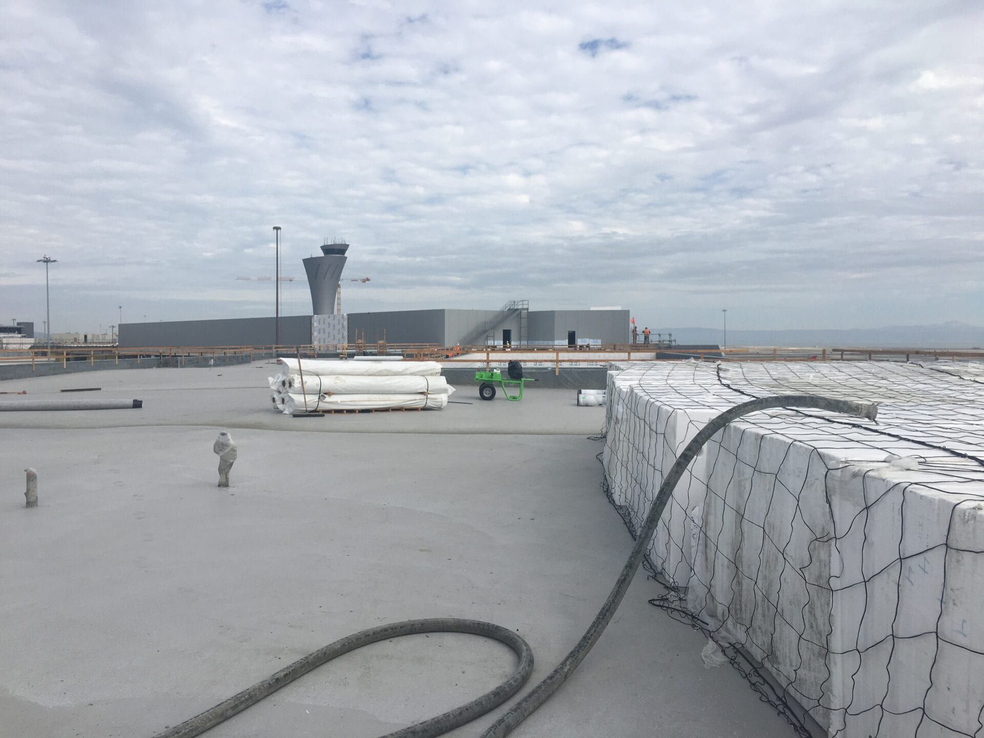 A construction site rooftop with white blocks in a wire cage, a gray hose, and an airport control tower in the distance.