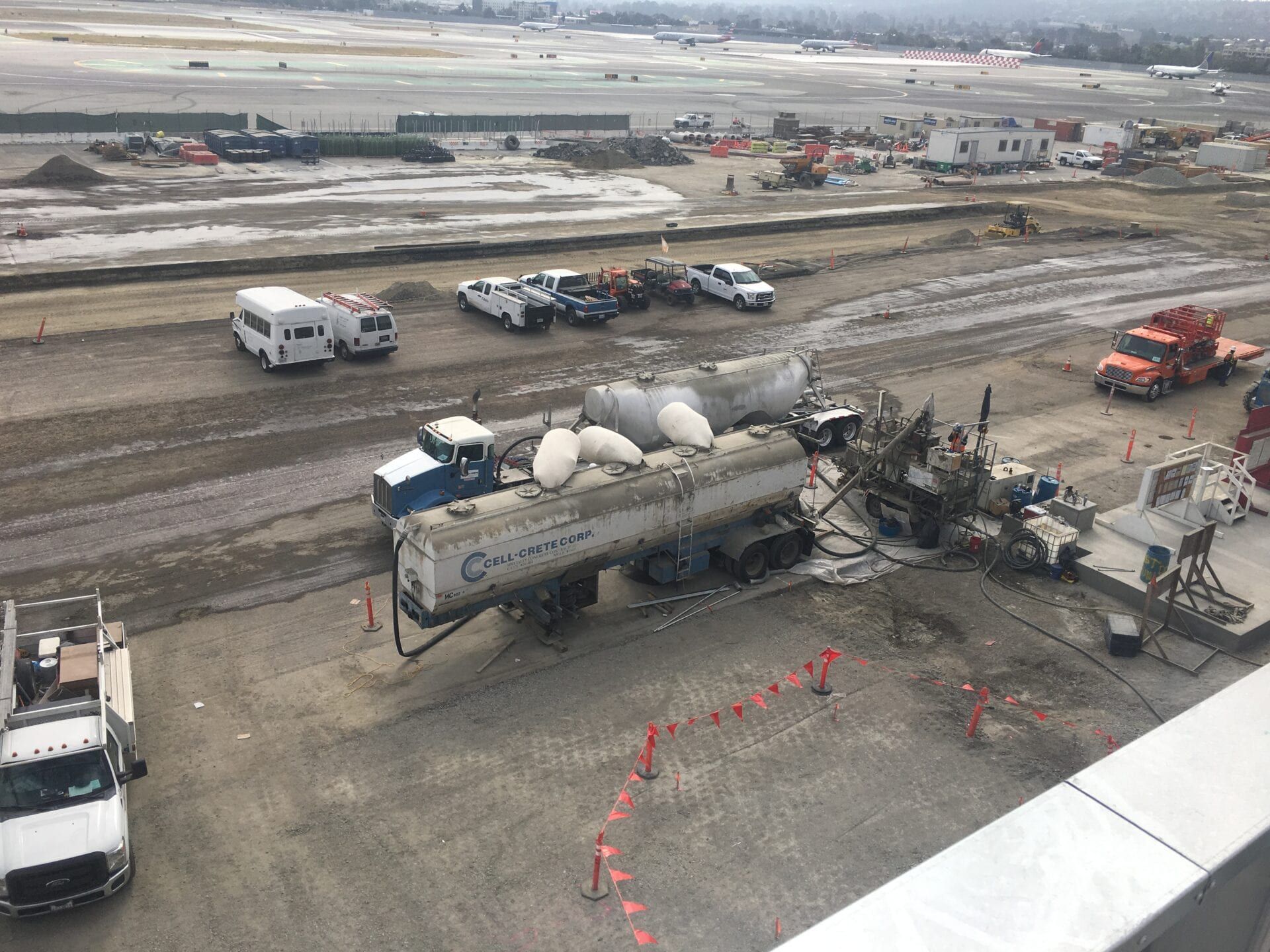 Construction site viewed from above, featuring large tankers, trucks, and equipment parked on a dirt lot near an airfield.