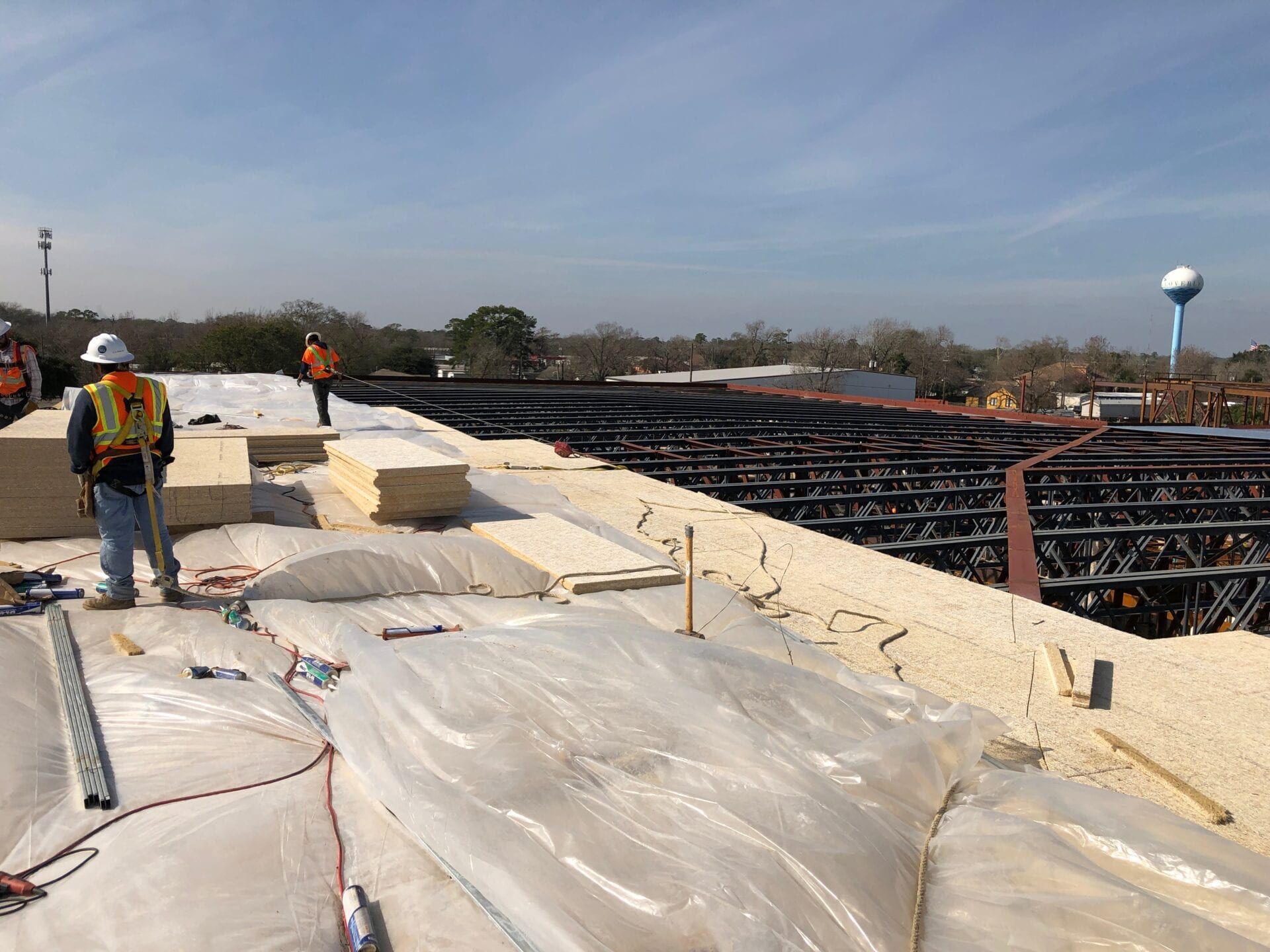 Two construction workers on a roof installing insulation boards over a metal frame under a clear blue sky.
