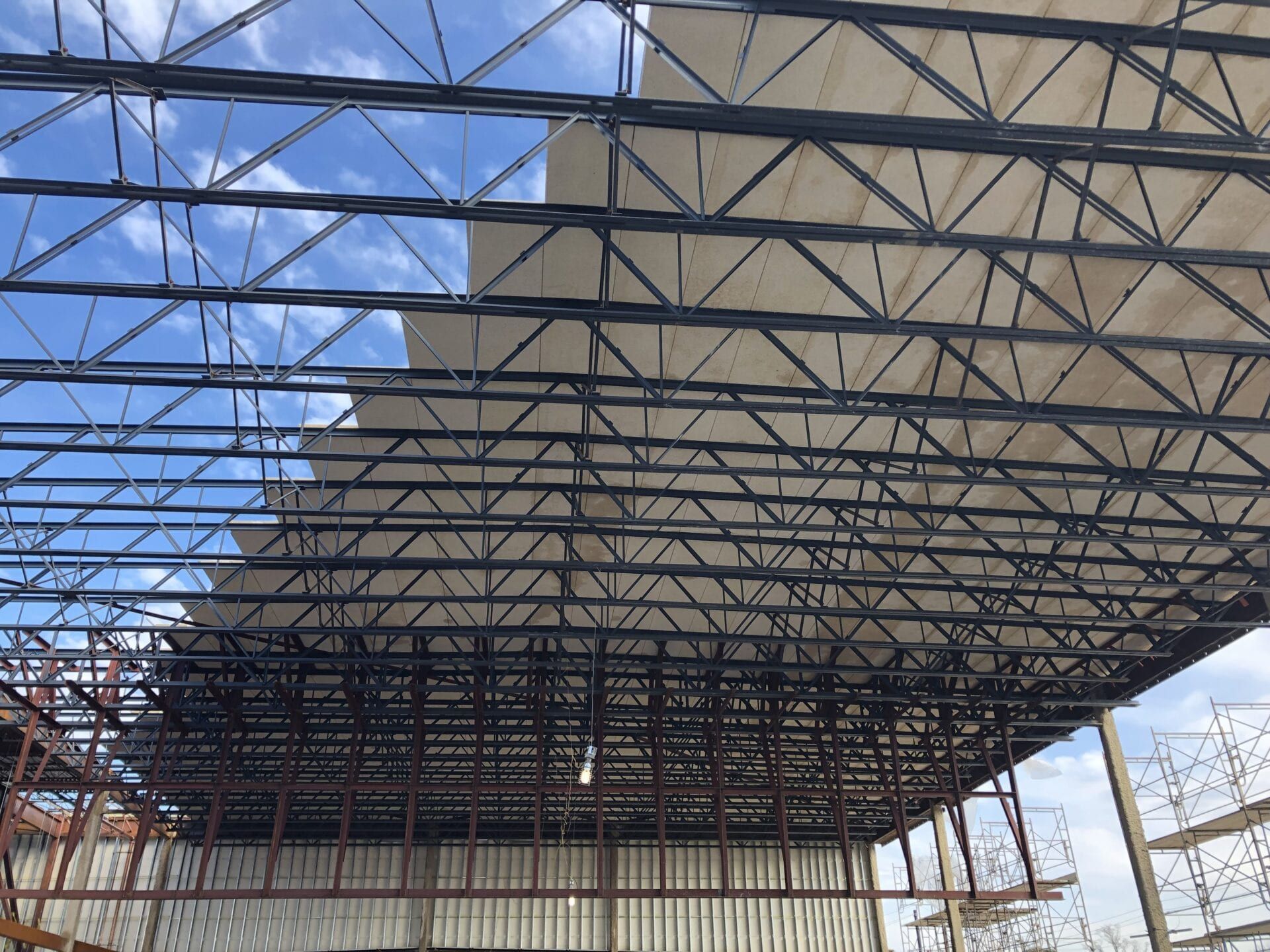 Exposed steel truss framework under construction against a partly cloudy blue sky.