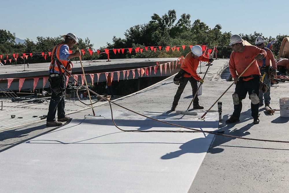Workers in safety gear finish a concrete surface on an outdoor construction site with orange flags in the background.