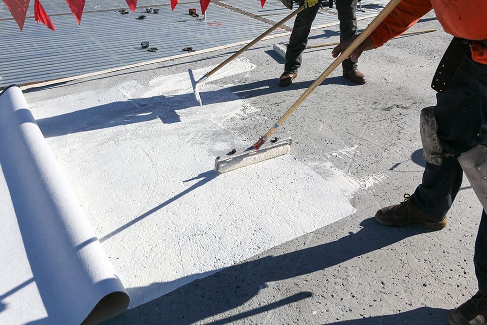 Construction workers spread white roof coating with long-handled rollers on a flat rooftop.