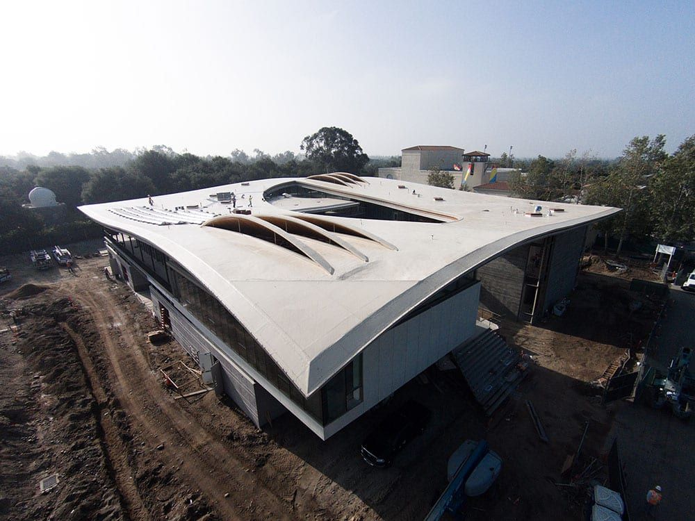 Aerial view of a modern building with a curved, light-colored roof and large skylights, under construction in a park.