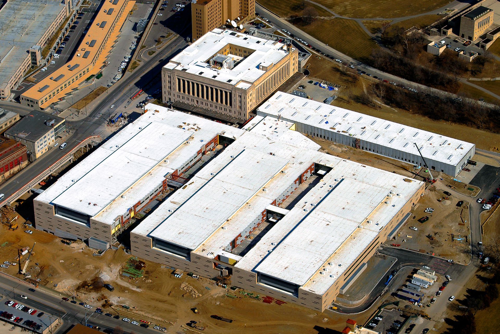 An aerial view of a large complex of multi-story buildings with white roofs, surrounded by construction and paved roads.