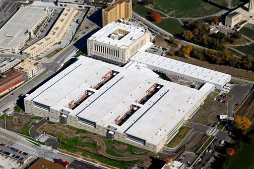 Aerial view of a large complex of connected buildings with white flat roofs, surrounded by parking lots and landscaping.