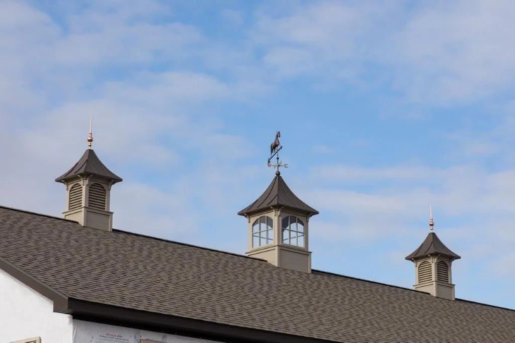 Barn roof with three cupolas, gray roof, light blue sky with clouds. A bird weathervane tops the center cupola.