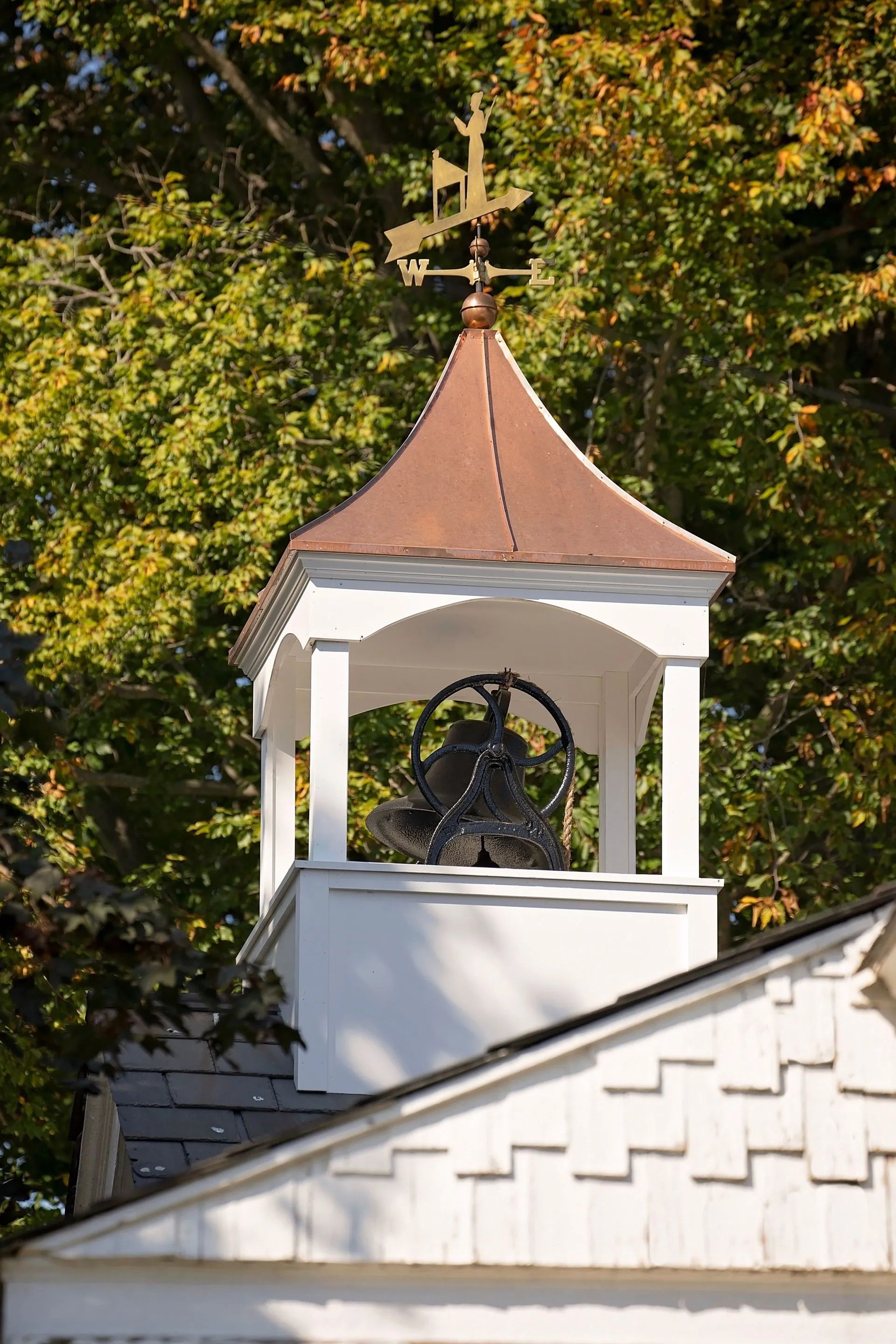 Bell tower with white structure, copper roof, and weather vane, set against leafy trees.