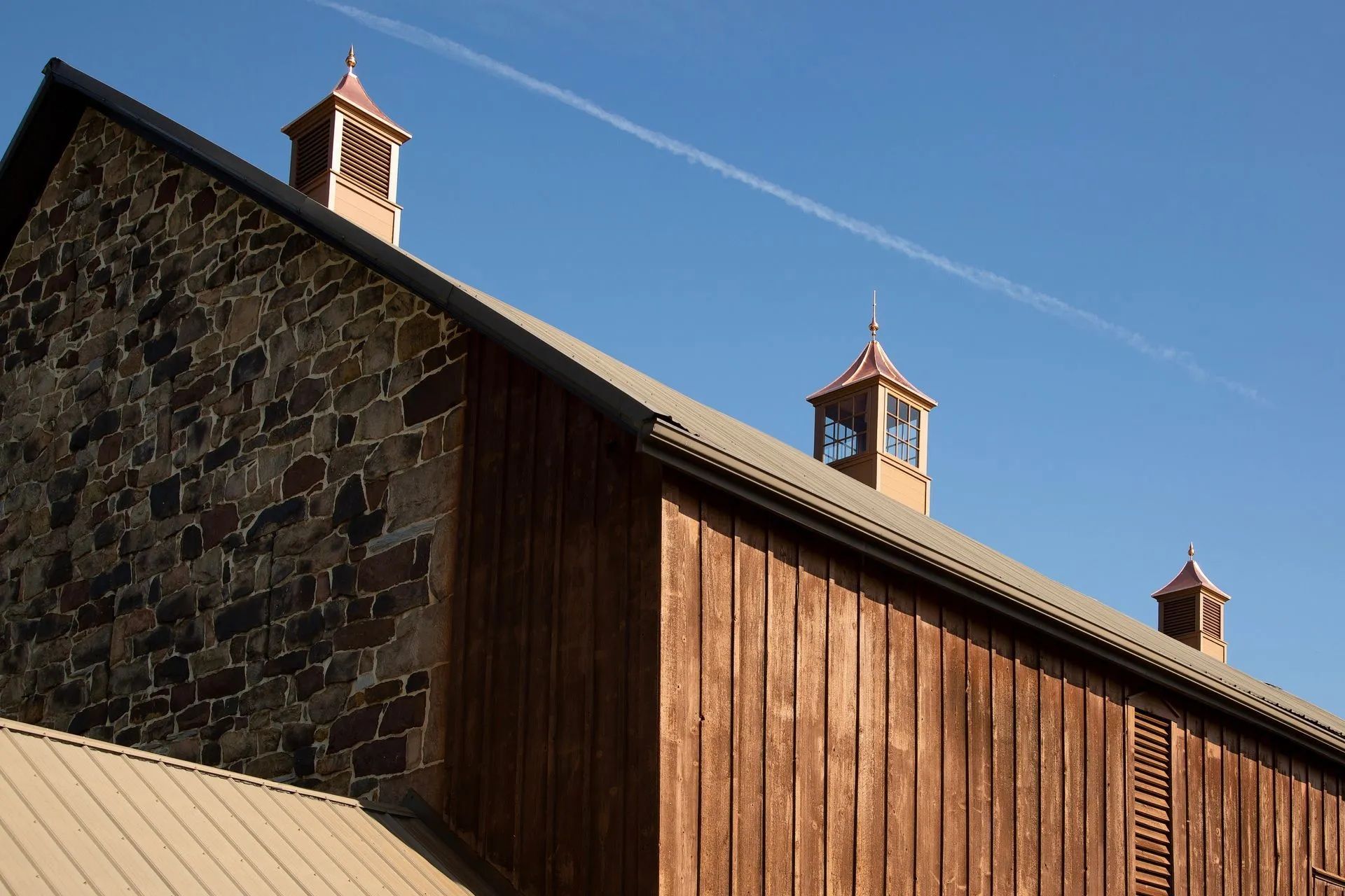 Barn with stone and wood siding, copper-colored roof and cupolas, against a clear blue sky.