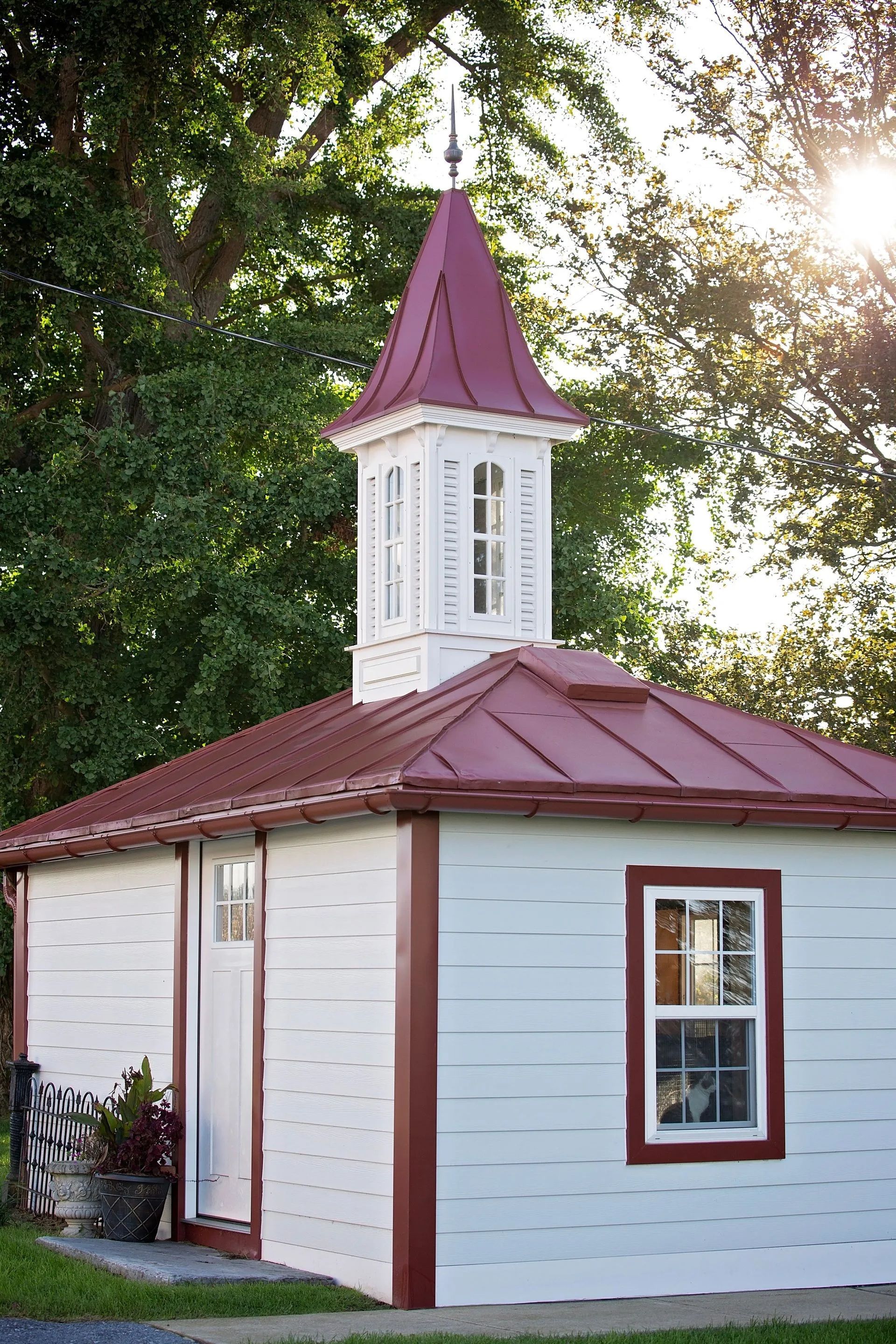 White and burgundy shed with tower, metal roof, and window. Sunlight and trees in background.